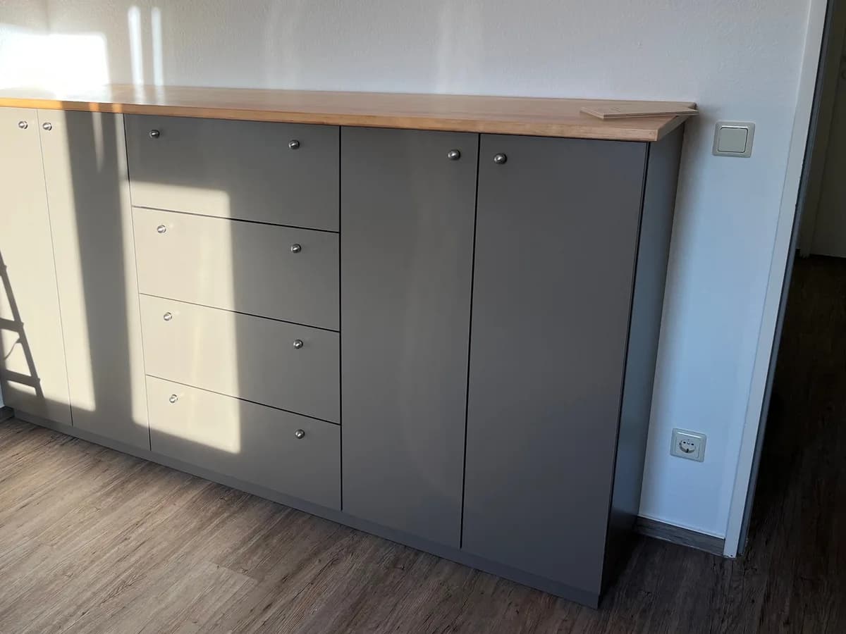 Sideboard mit Fronten in Taupe: in der Mitte vier Schubkästen, links und rechts je zwei Türen mit kleinen runden Knöpfen, darüber eine durchgehende Deckplatte aus geölter massiver Erle. Die Sonne wirft Streifen auf die Fronten.