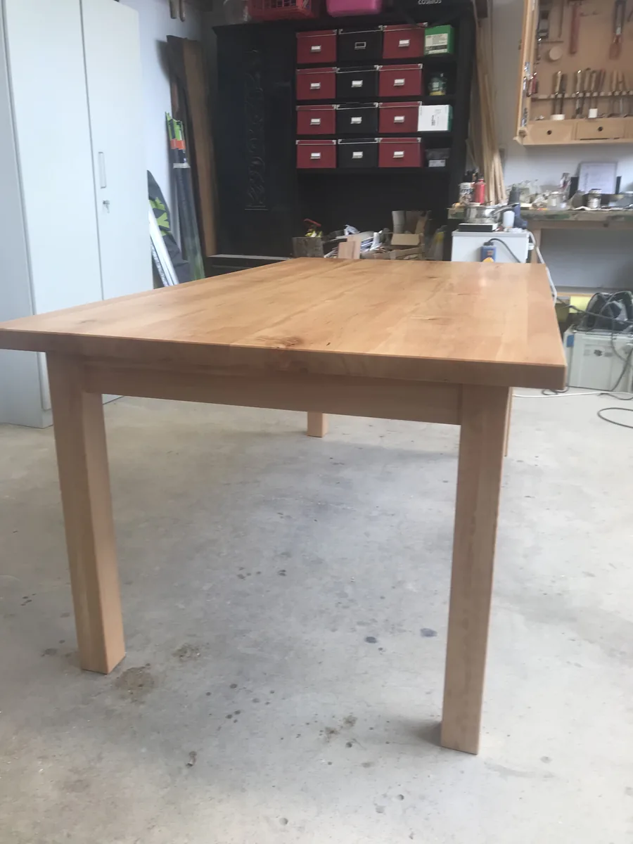 The table from the end: the apron beneath the top, the legs flush at the corners, the workshop tool wall behind.