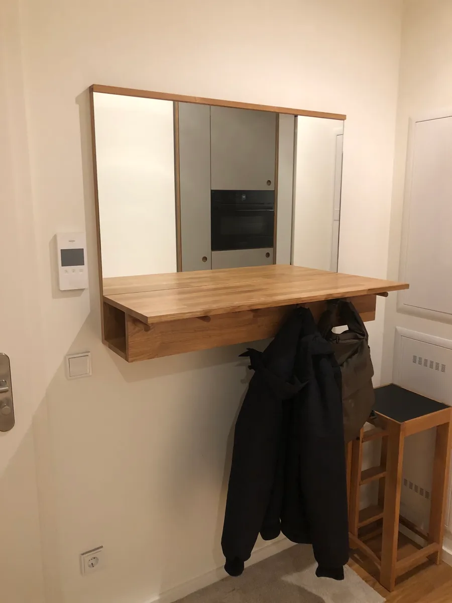 The oiled oak hallway unit from the side: beneath the projecting shelf the row of hooks, with two dark jackets and a bag hanging from them, the kitchen opposite reflected in the mirror.