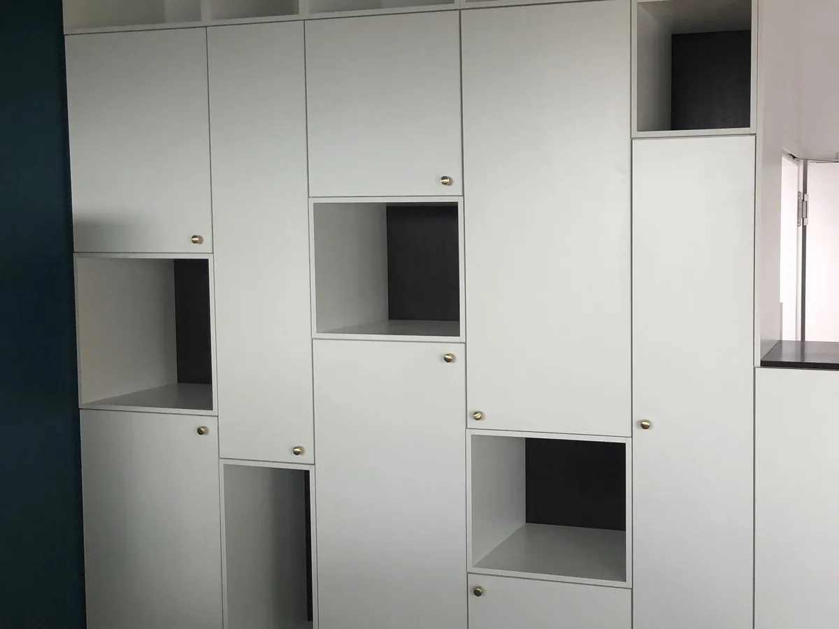 Weiße Schrankwand mit wechselnd geschlossenen Türen und offenen Fächern, die Fächer innen dunkel abgesetzt. Kleine runde Knöpfe, links eine dunkelgrüne Wand, rechts ein Fenster.
