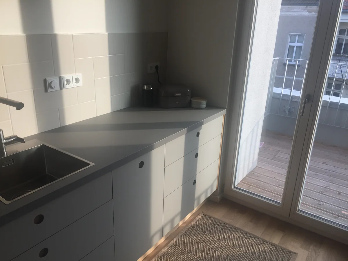 The end of the kitchen run in daylight: a stainless-steel sink in the grey worktop, handle-free fronts with round finger pulls, the balcony door on the right. Sunlight throws stripes across the tiles.