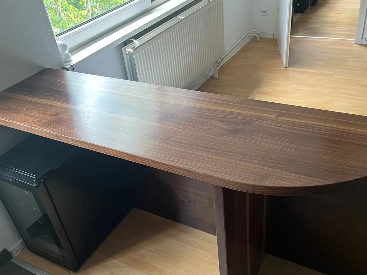 Gebogene Platte aus massivem Nussbaum zwischen Küche und Wohnraum, auf dunklem Wangengestell, die Kante läuft abgerundet durch.