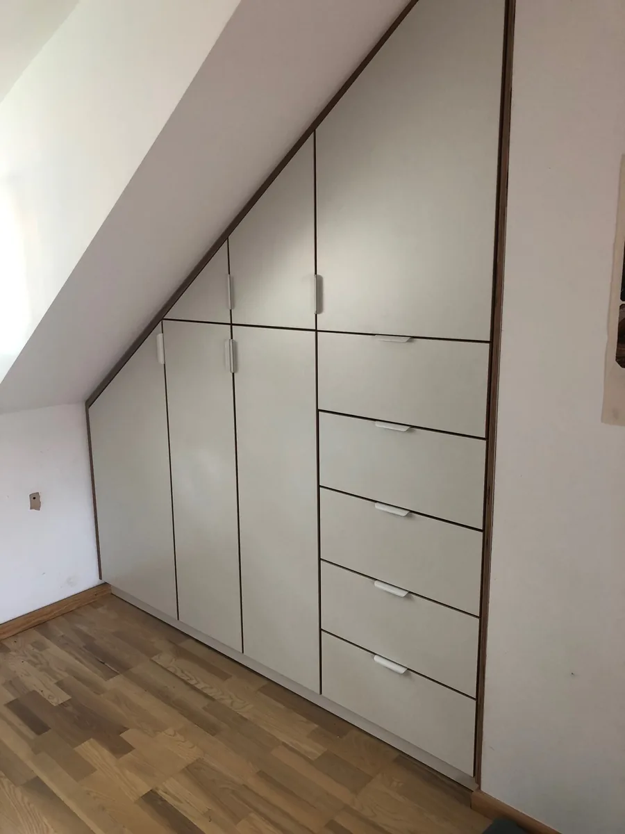 Der fertige Einbauschrank unter der Dachschräge: weiße Fronten mit Nussbaum-Kanten, Türen und Schubladen auf den Winkel der Schräge zugeschnitten, die Griffe montiert.