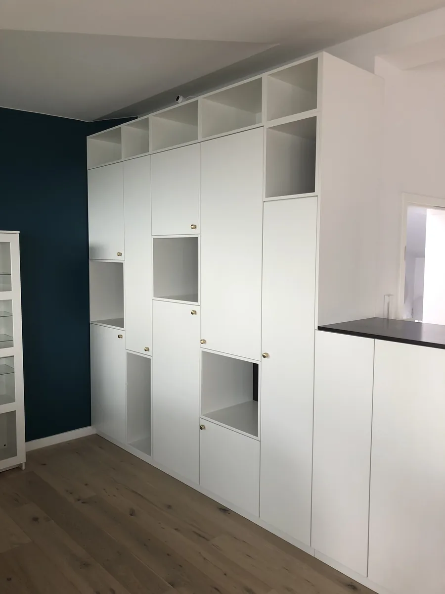 Die ganze Schrankwand im Raum: raumhohe weiße Fronten mit kleinen Messingknöpfen, dazwischen offene Fächer, oben eine Reihe offener Ablagen bis zur Decke. Links eine dunkelgrüne Wand und ein Vitrinenschrank, rechts der niedrigere Teil mit dunkler Platte, der den Raum teilt, unten Eichendielen.