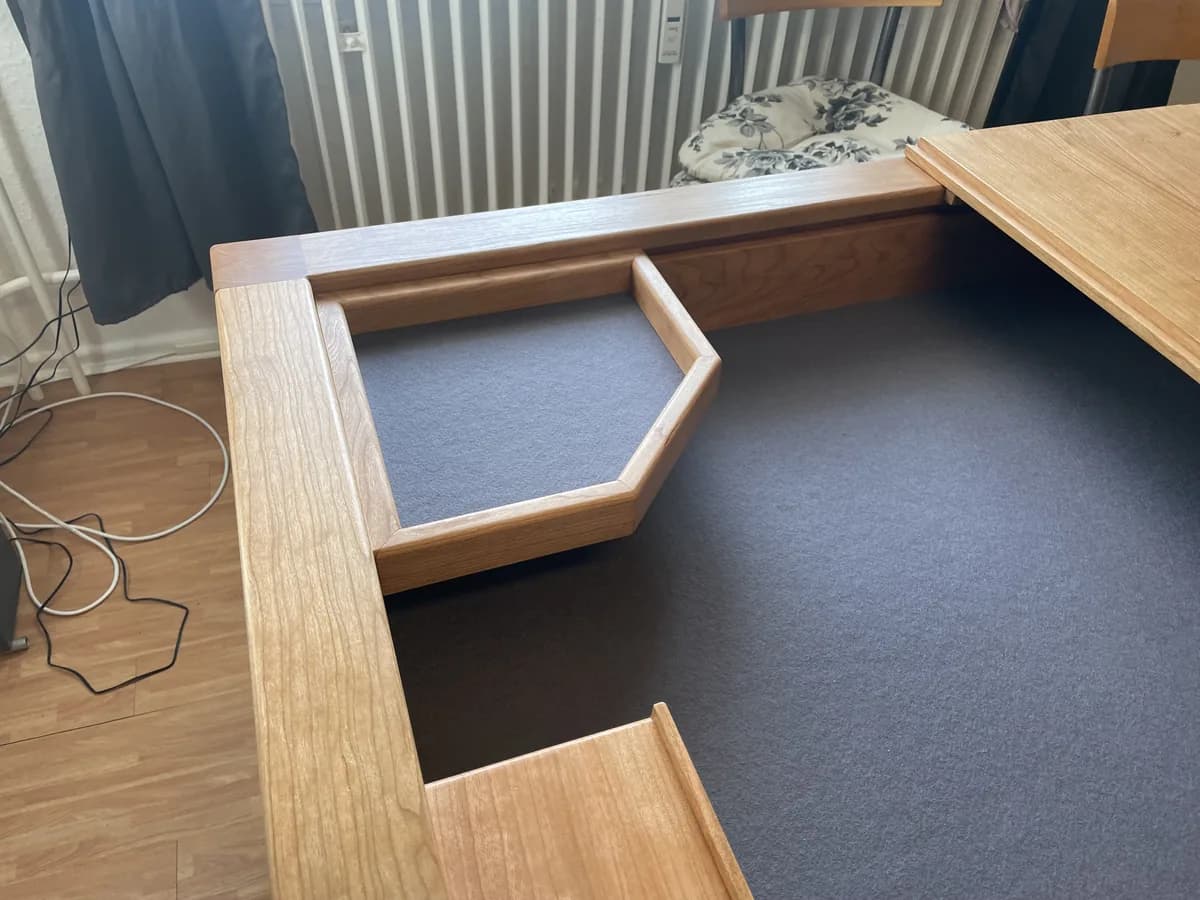 One corner of the games table close up: the five-sided cherry dice tray, lined with felt, hooked into the groove along the edge. A radiator and curtain behind.