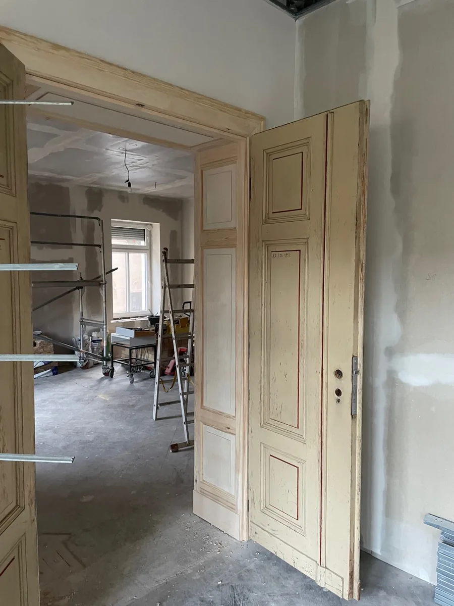 The same double doors open, from the side: the new panelled lining in the opening, one leaf with its old paint and red-lined mouldings. The room beyond under renovation, with scaffold and ladder.