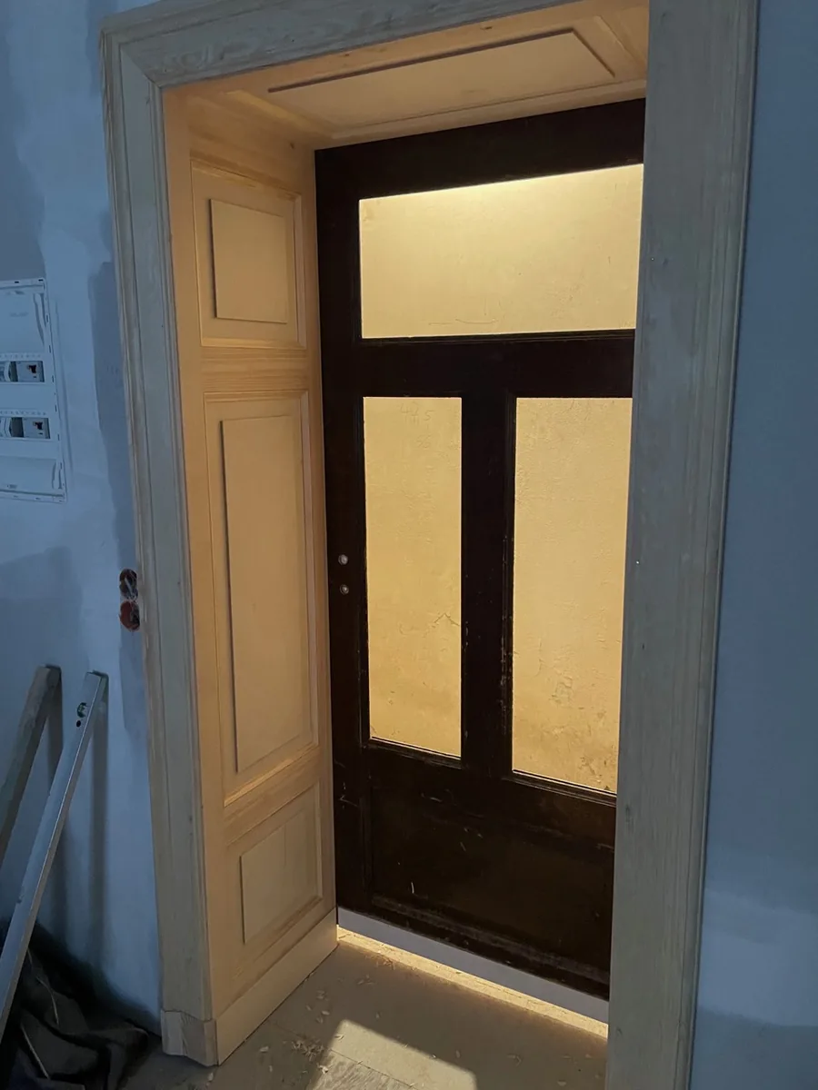 A second door frame with panelled lining and architrave, holding a dark glazed door with glazing bars; warm light comes through the glass from the next room. A fuse box on the bare wall to the left.