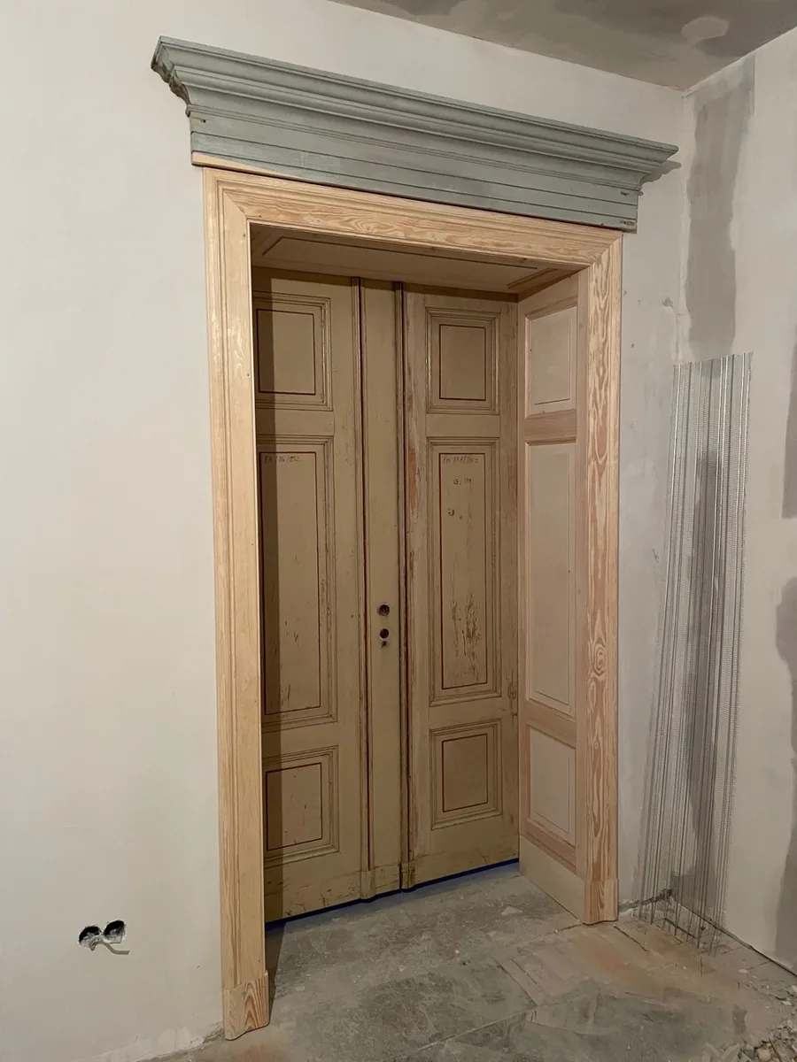 New pine door frame with panelled architrave and the restored crowning above; in it the old two-leaf door with its old paint. Skimmed walls, a plaster bead to the right, before painting.