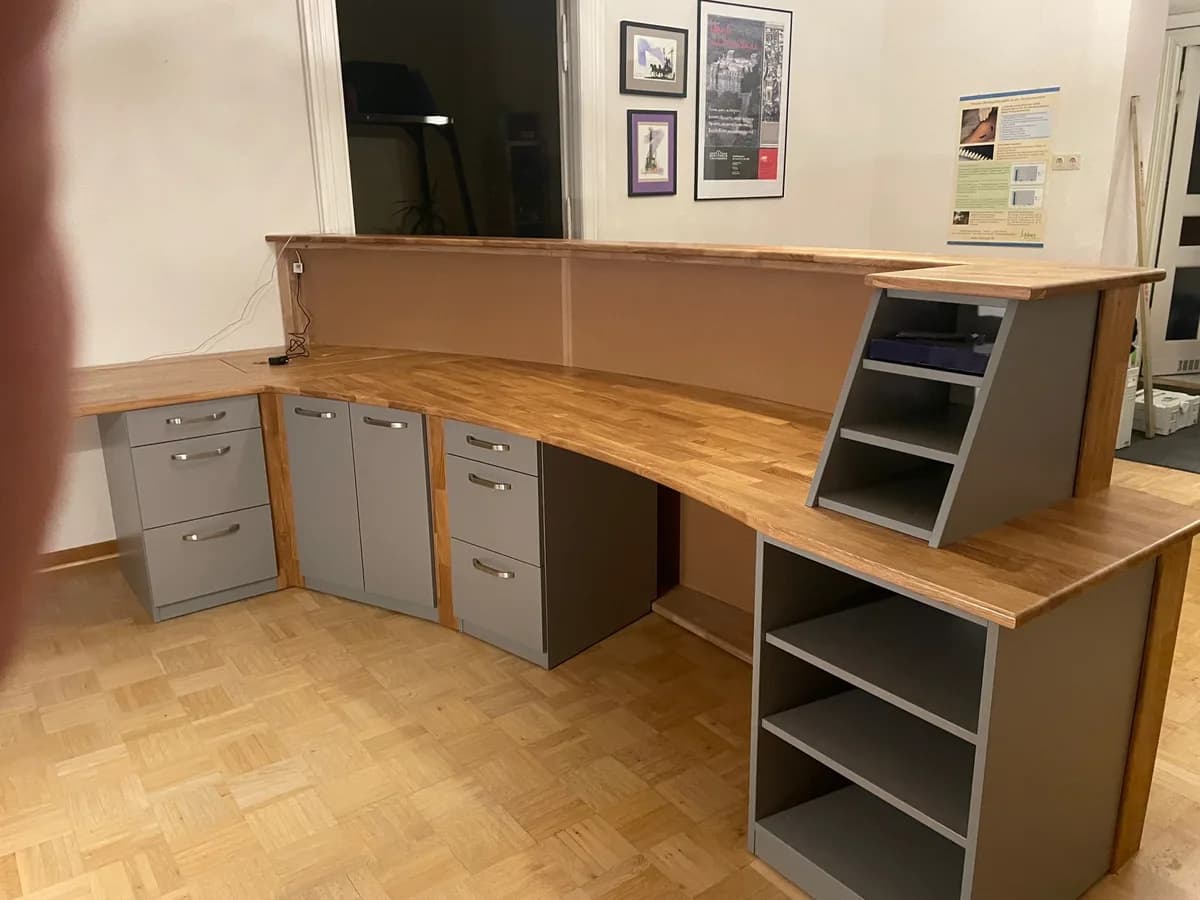 Der Tresen von der Arbeitsseite: eine durchgehende Massivholzplatte über grauen Unterschränken mit Schubladen und Türen, rechts ein offenes Regal unter der erhöhten Theke, an der Wand gerahmte Bilder.