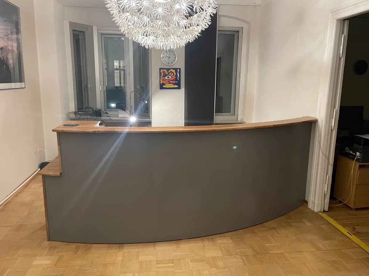 Der Tresen von vorn: die graue, über einen großen Radius gebogene Front mit Massivholzplatte, darüber eine große weiße Kugelleuchte, dahinter Fenster und eine Wanduhr.