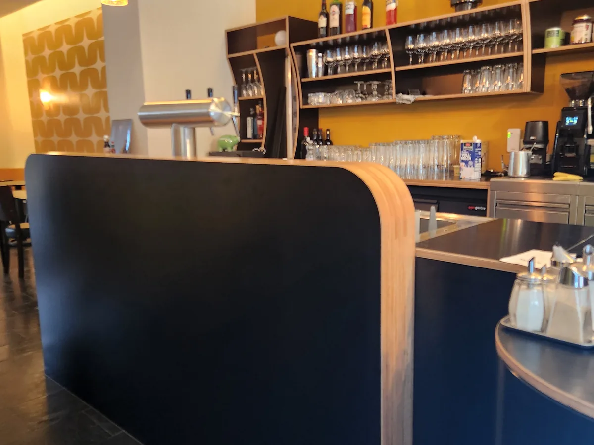 The rounded corner of the counter close up: a black work surface with the oiled striped plywood edge carried around it, and behind it the tap, the glass shelves and the coffee machine.