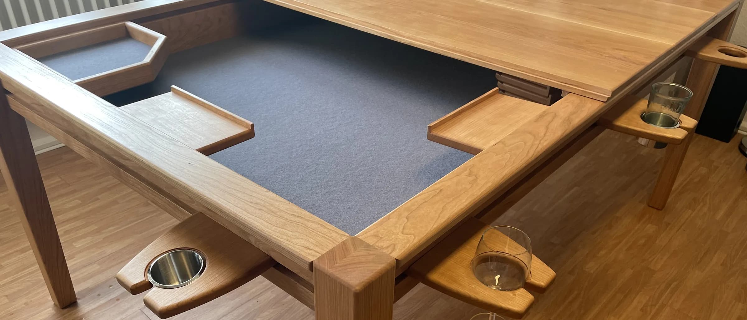 Spieltisch aus Kirschbaum: die vertiefte Spielfläche mit dunkelgrauem Filz, zur Hälfte von einer aufgelegten Massivholzplatte bedeckt. An den Rändern eingehängte Glashalter mit Gläsern und mehrere Ablageschalen.