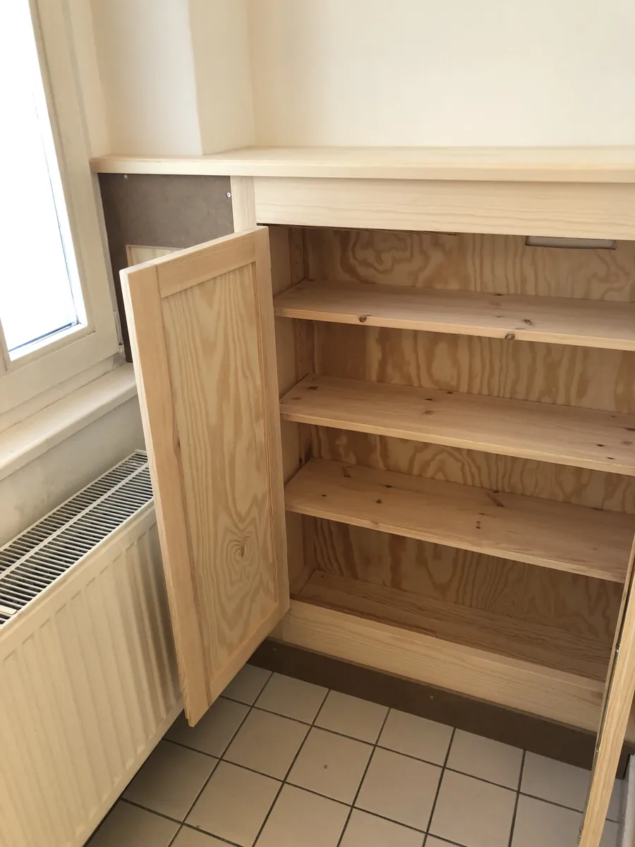 Der Einbauschrank aus Kiefer mit einer geöffneten Rahmentür: dahinter vier Böden aus Kiefer, die Rückwand aus Kiefernsperrholz, links das Fenster mit Heizkörper, unten weiße Bodenfliesen.