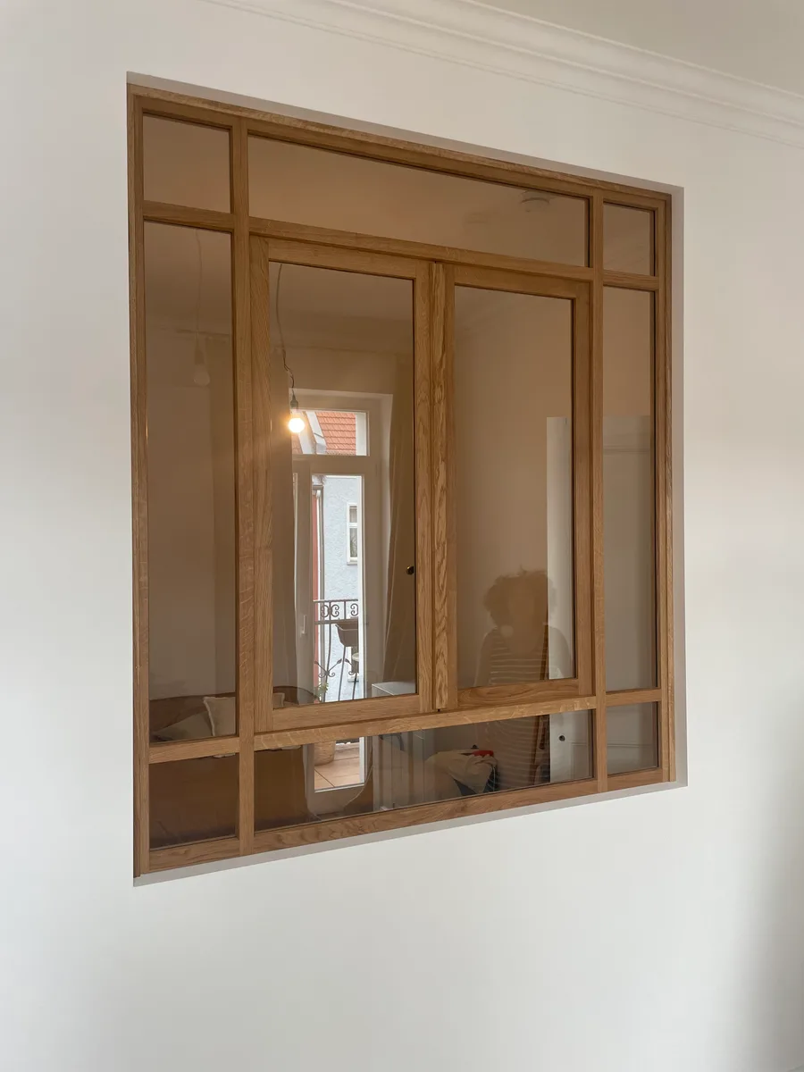 The closed oak interior window in the white wall: two casements with a handle, fixed panes above and to the sides. Through the glass the adjoining room with a pendant lamp, the balcony door and a dressmaker’s dummy.
