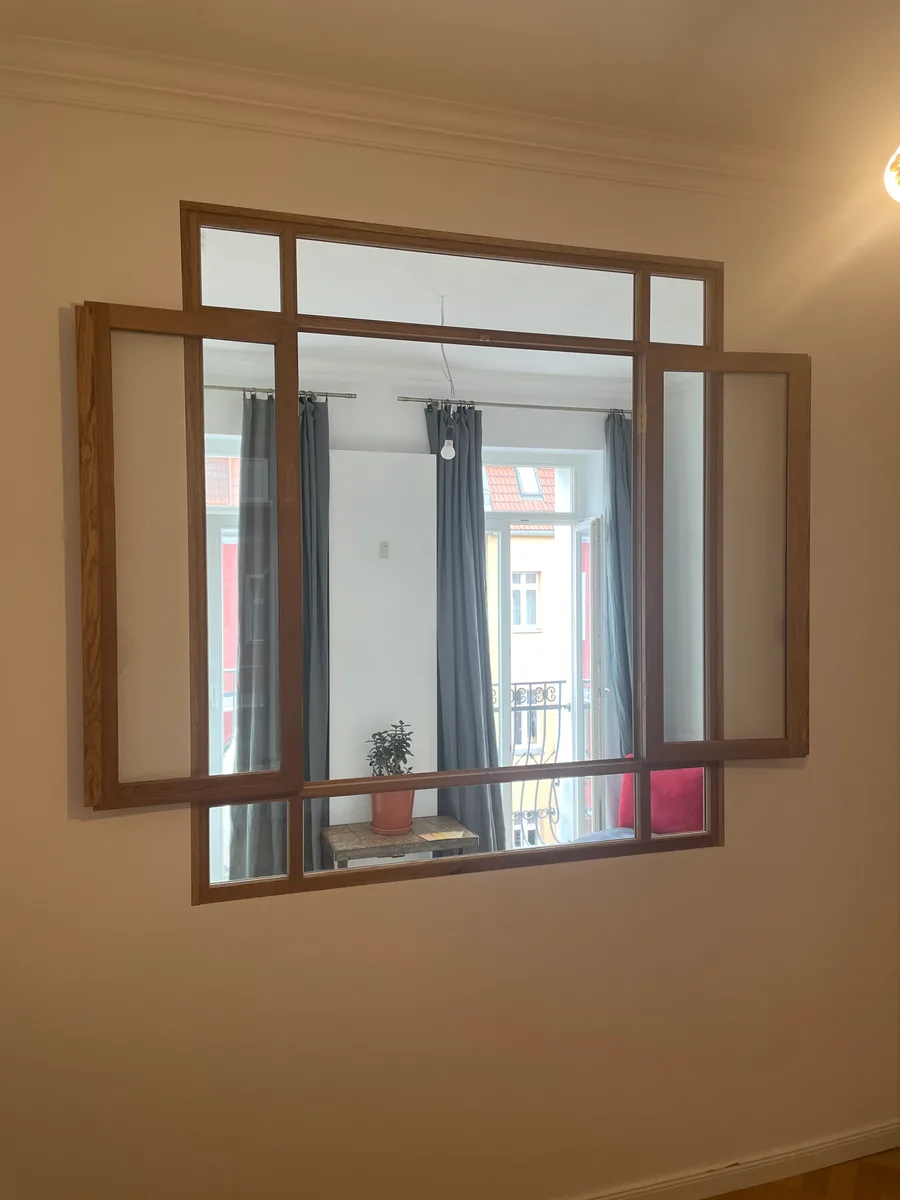 The oak interior window with both casements open, seen from the inner room: beyond it the outer room with grey curtains, the balcony door and a pot plant on a small table.