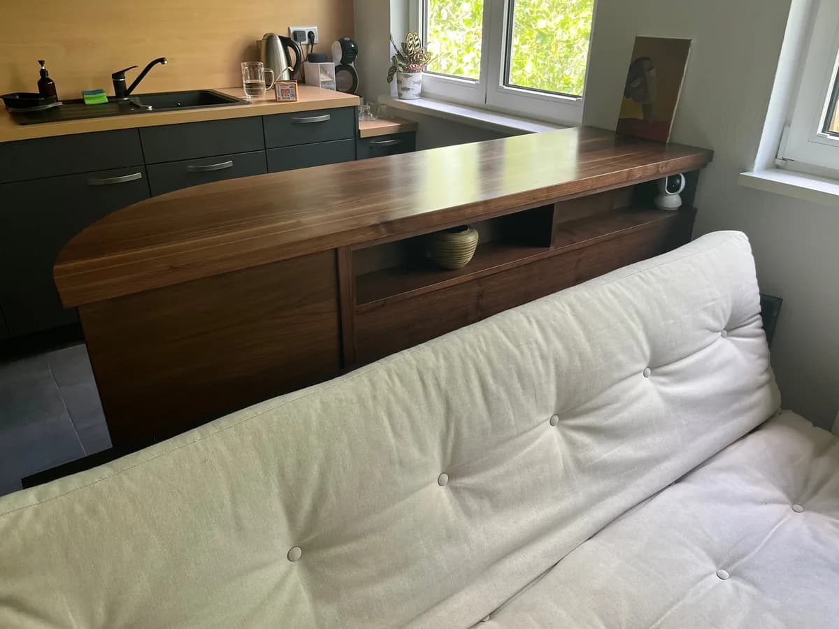 Die gerundete Nussbaumplatte vom Wohnraum aus gesehen: dahinter die Küchenzeile, davor ein Sofa. Unter der Platte ein offenes Fach.