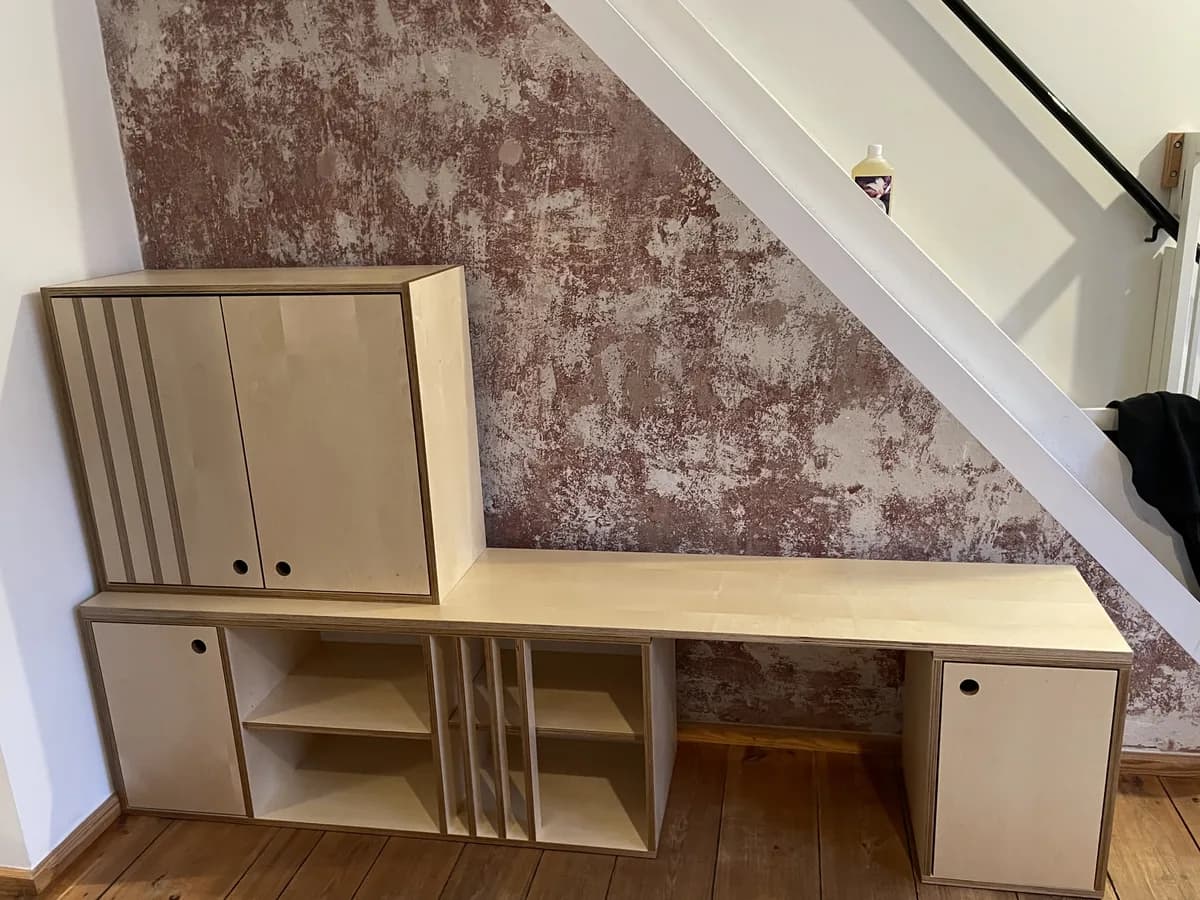 Sideboard aus hellem Ahorn-Multiplex unter einer Treppe: unten offene Fächer und Türen, darüber eine durchgehende Platte, die rechts zum Schreibtischplatz wird, links ein aufgesetzter geschlossener Korpus mit zwei Türen für Fernseher und Stereoanlage. Dahinter eine Wand in rot-grauer Spachteltechnik.
