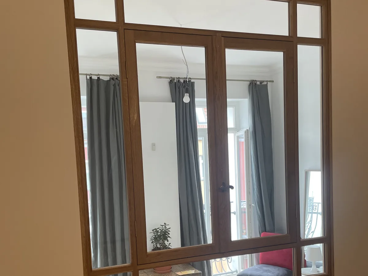 Innenfenster mit Rahmen aus Eiche, vom inneren Zimmer aus gesehen: zwei Flügel mit Griff, darüber und seitlich feste Scheiben. Durch das Glas fällt Tageslicht aus dem Nebenraum mit Balkontür und grauen Vorhängen.