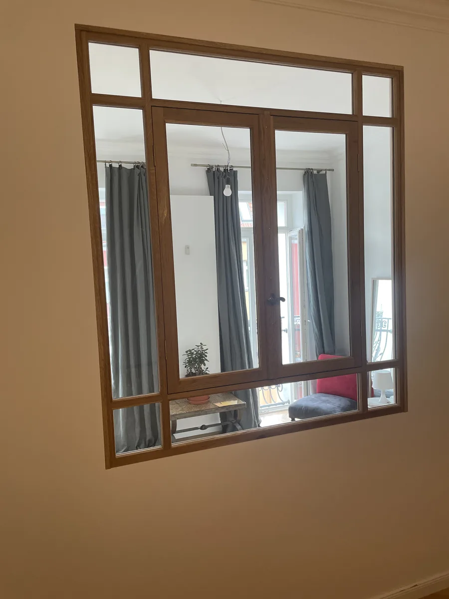 Interior window framed in oak, seen from the inner room: two opening casements with a handle, fixed panes above and to the sides. Daylight comes through the glass from the adjoining room with its balcony door and grey curtains.