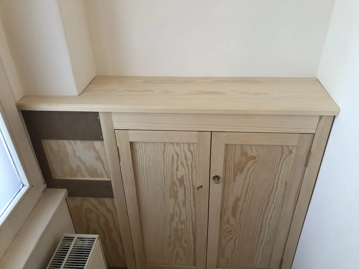 Einbauschrank aus Kiefer in einer Nische: zwei Rahmentüren mit Füllung und einem Schlüsselschild, darüber eine durchgehende Deckplatte, links das Fenster mit Heizkörper.