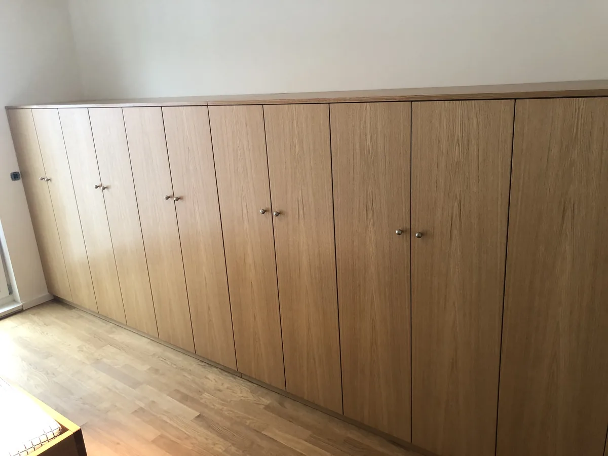 Langer Schlafzimmerschrank in Eiche natur, etwa hüfthoch bis brusthoch, über die ganze Wand geführt. Neun Türen mit kleinen runden Knöpfen, davor Parkett.