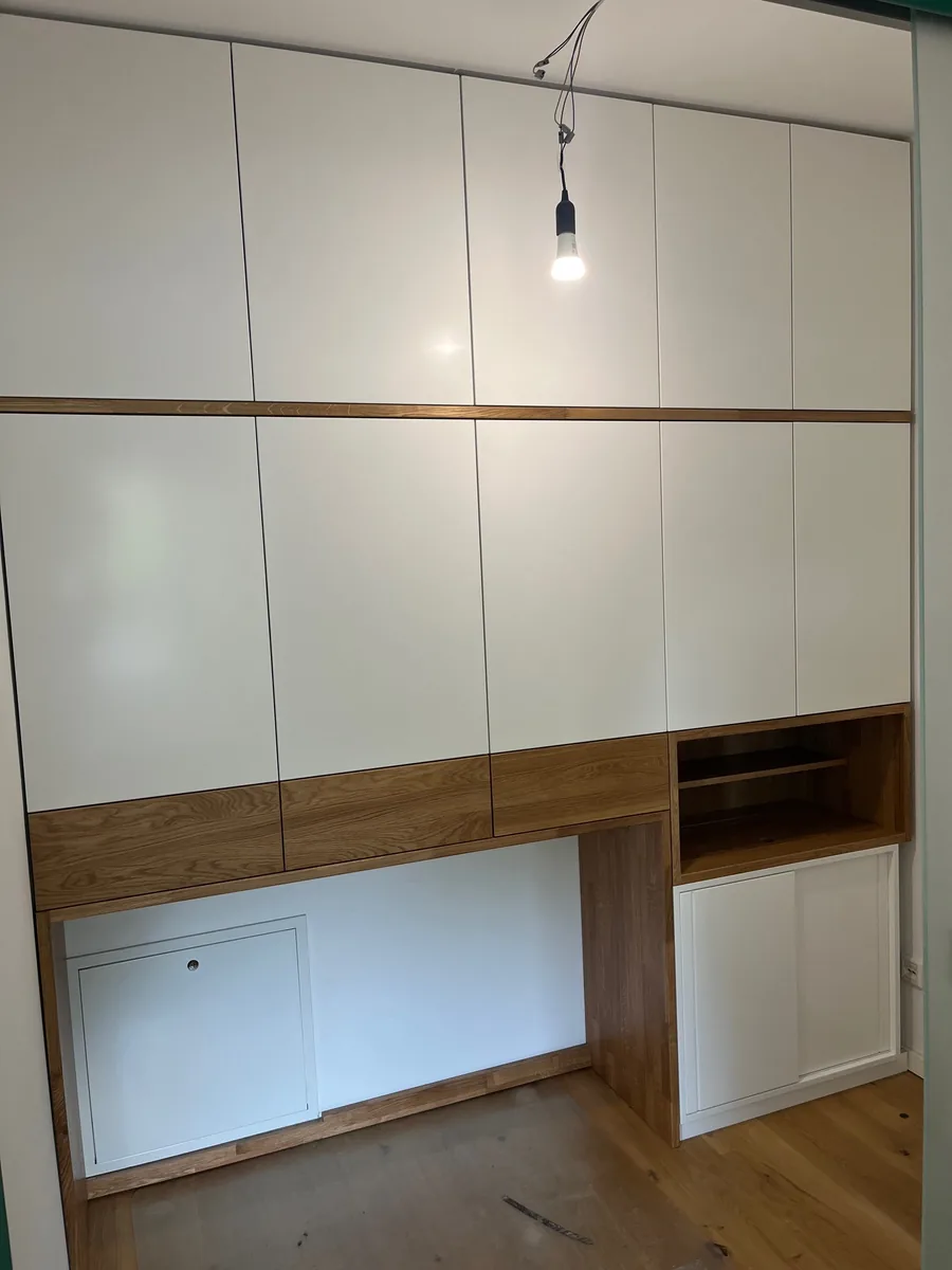 Room-height fitted wall in white with a continuous band of solid oak, six doors in the upper section and an open compartment above the workspace on the right.
