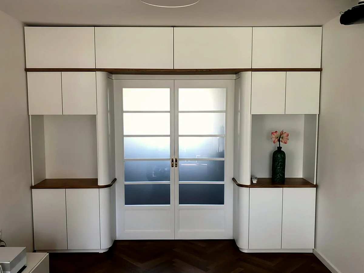 Raumhoher Einbauschrank um eine zweiflügelige Glastür, mit offenen Nischen und Ablagen aus Eiche. Die senkrechten Kanten sind gerundet.