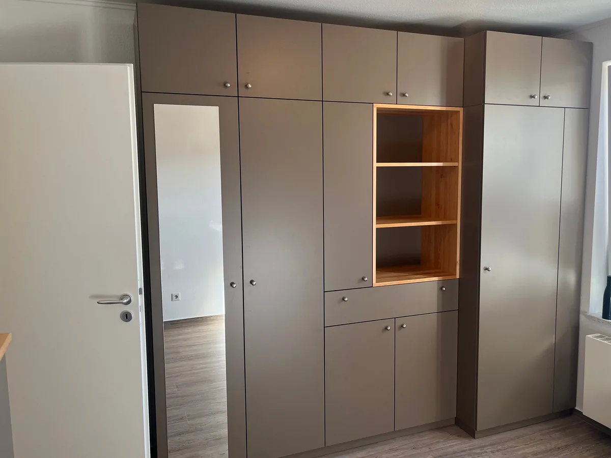Raumhohe Einbauschrankwand mit Fronten in Taupe, einer Spiegeltür links und einem offenen Regalfach aus Erle in der Mitte, rechts ein Versprung zum Fenster hin. Links die Zimmertür, rechts das Fenster mit Heizkörper.