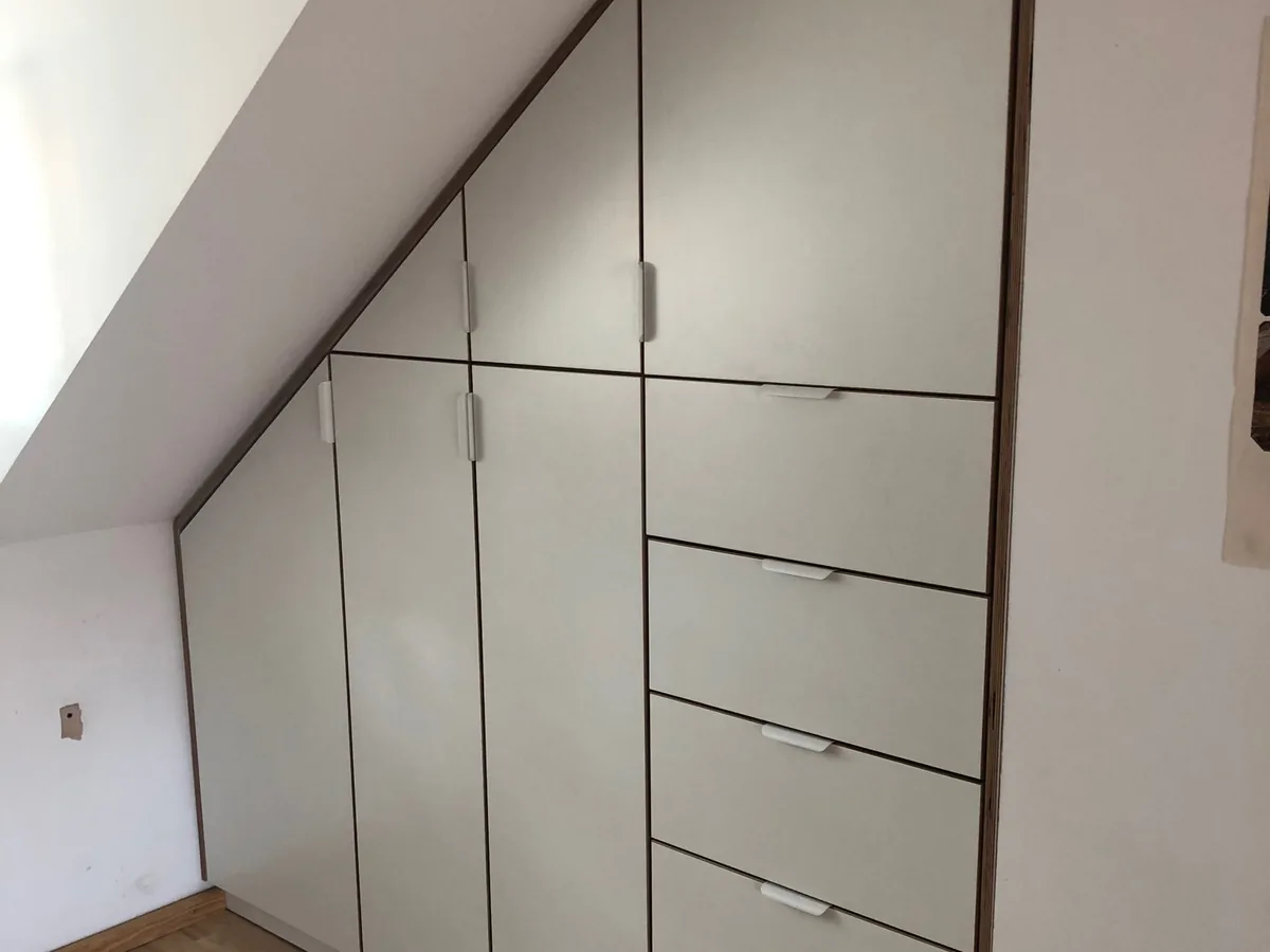 Der fertige Einbauschrank unter der Dachschräge: weiße Fronten mit Nussbaum-Kanten, Türen und Schubladen auf den Winkel der Schräge zugeschnitten, die Griffe montiert.