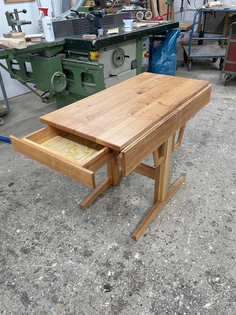 Die kurze Seite des Tisches, die Sitzseite: der flache Schubkasten herausgezogen, Seiten und Front aus Massivholz, der Boden aus hellem Sperrholz, dahinter das Untergestell auf Kufen.
