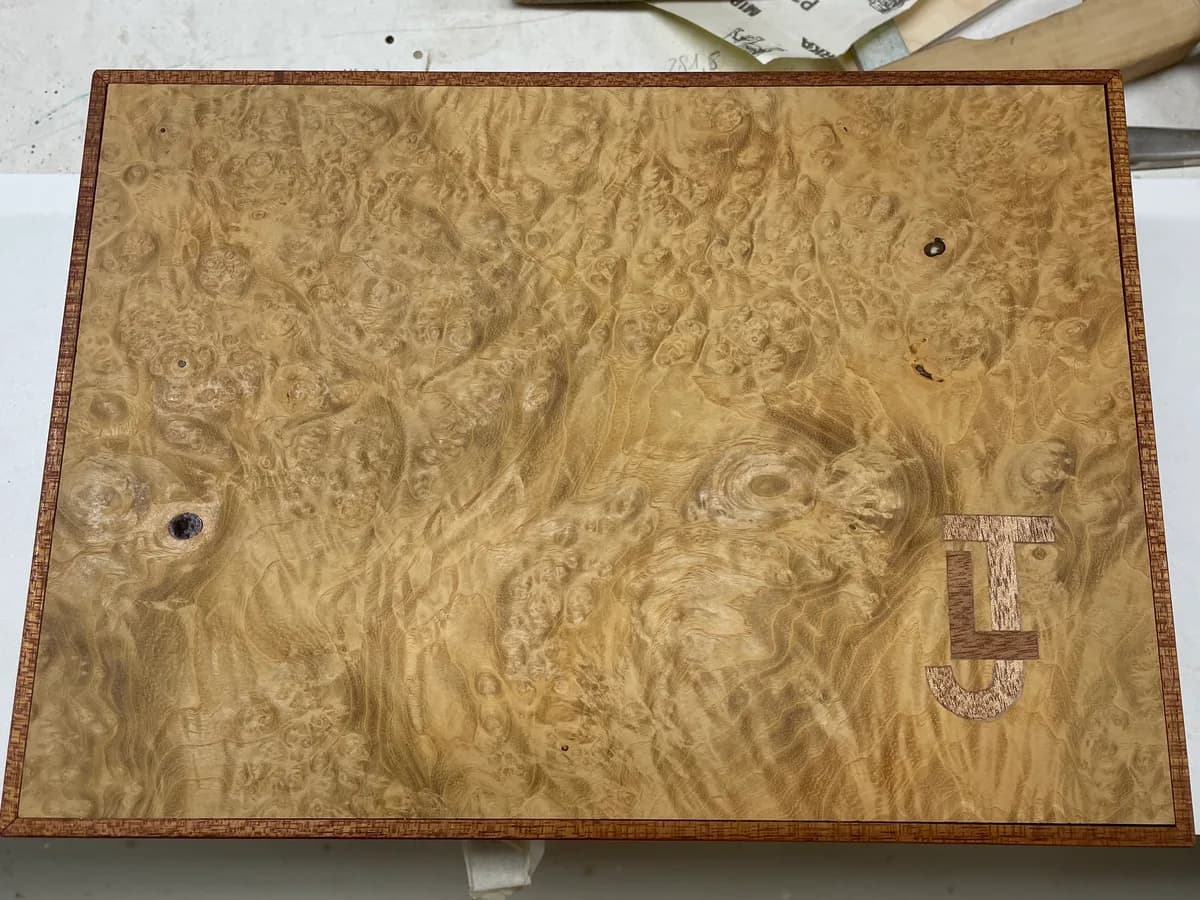 The lid of the box from above, on the workbench: the figured grain of the masur ash, a narrow mahogany border, and the inlaid mahogany initials at the lower right.