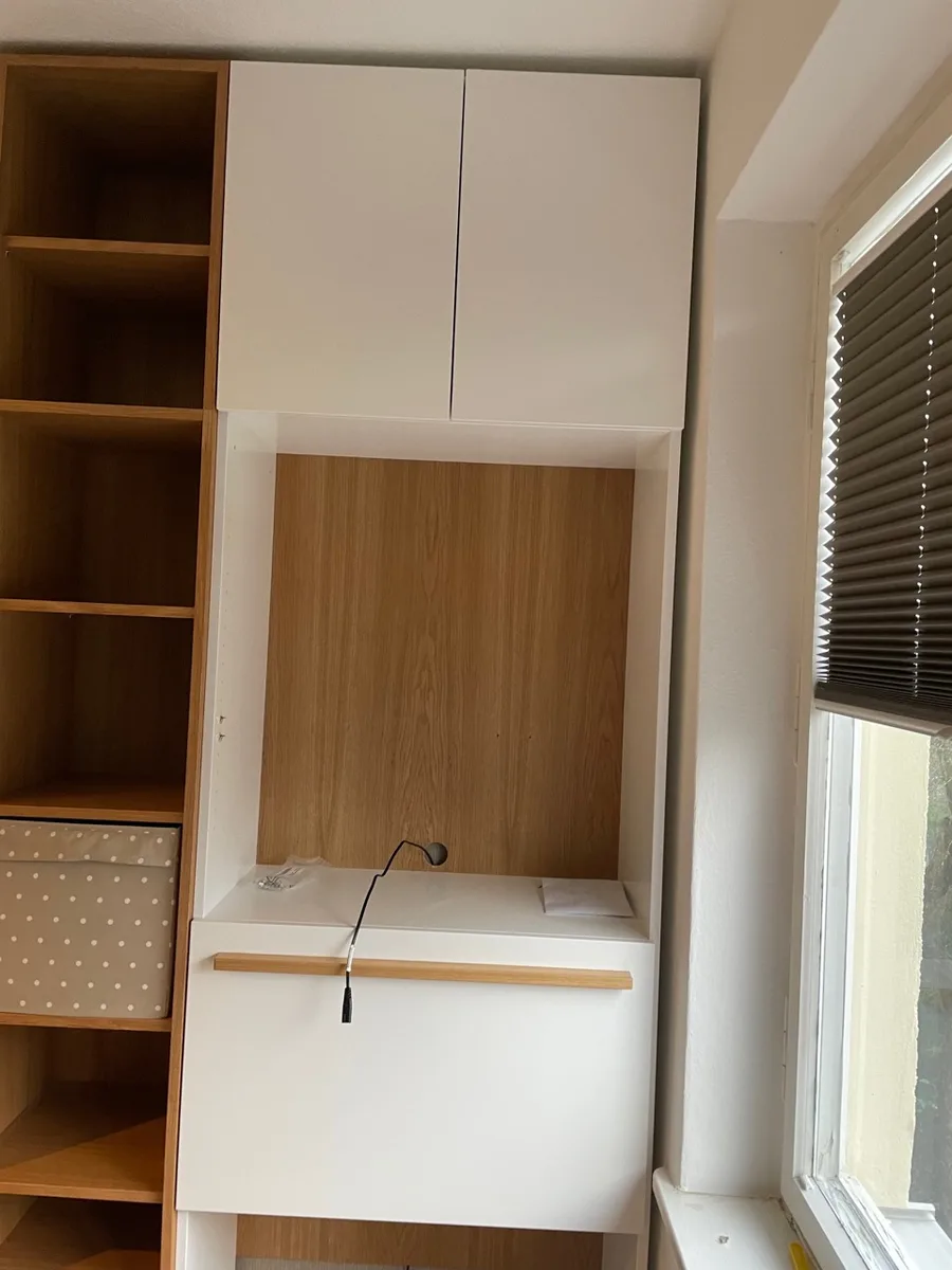 The writing desk by the window: a fold-down flap with a handle rail, the oak back panel with a small lamp behind it, two doors above; the shelving tower on the left, the window with a pleated blind on the right.