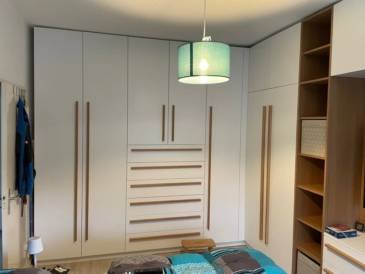 Raumhohe Schrankwand im Schlafzimmer: weiße Fronten mit langen senkrechten Griffleisten aus Eiche, in der Mitte sechs Schubladen, rechts der Anschluss an einen offenen Regalturm. Darüber eine türkise Pendelleuchte, unten das Bett.