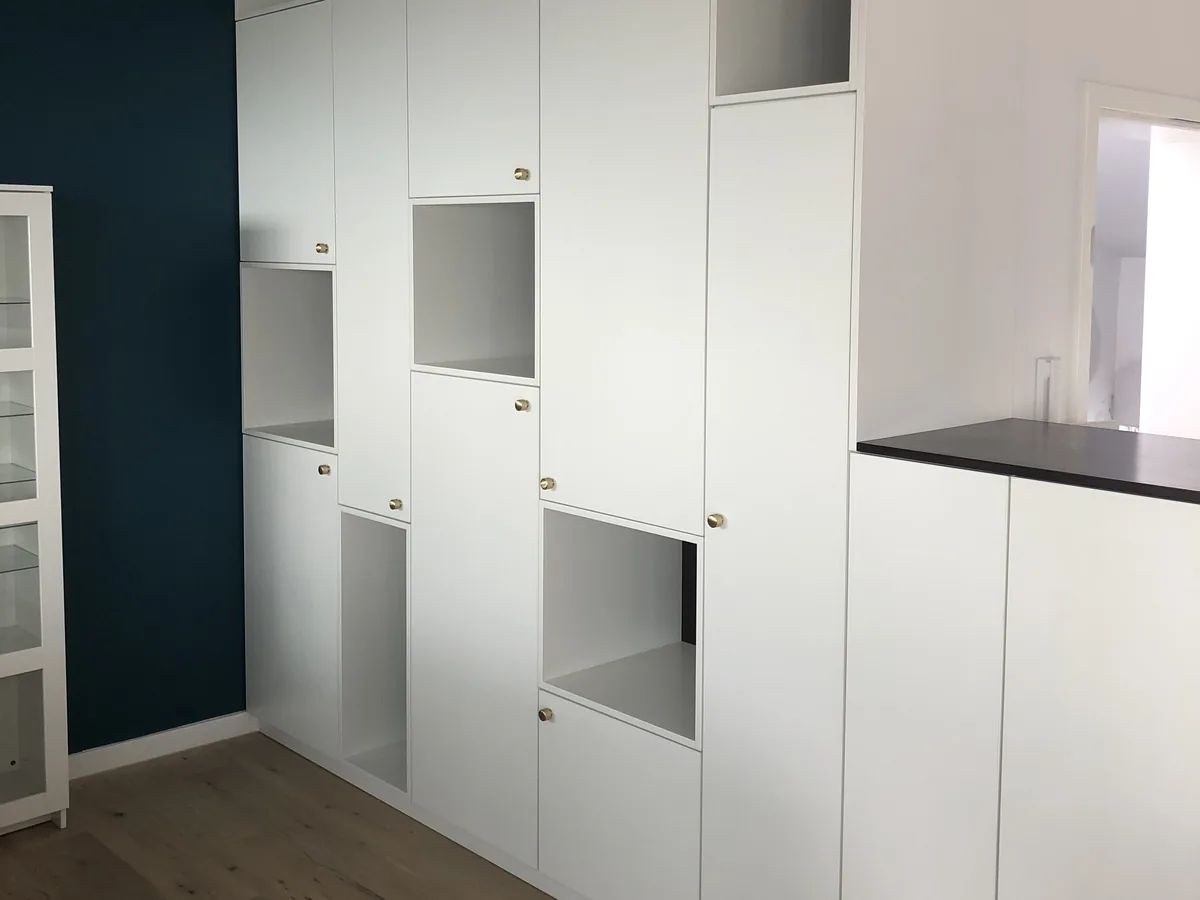 Die ganze Schrankwand im Raum: raumhohe weiße Fronten mit kleinen Messingknöpfen, dazwischen offene Fächer, oben eine Reihe offener Ablagen bis zur Decke. Links eine dunkelgrüne Wand und ein Vitrinenschrank, rechts der niedrigere Teil mit dunkler Platte, der den Raum teilt, unten Eichendielen.