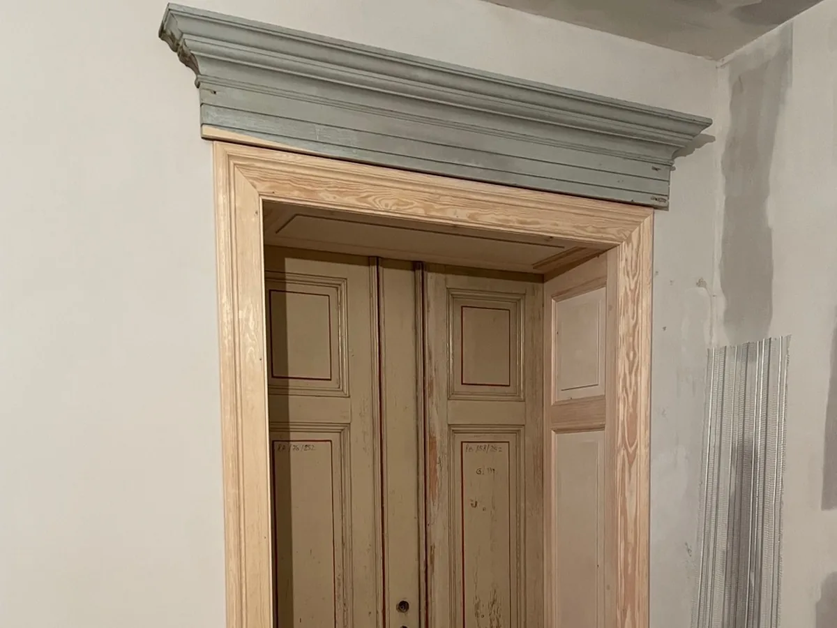 Neue Türzarge aus Kiefer mit Füllungen in der Bekleidung, darüber die restaurierte Bekrönung; darin die alte zweiflügelige Tür mit ihrer alten Farbfassung. Wände verspachtelt, rechts eine Putzschiene, vor dem Anstrich.
