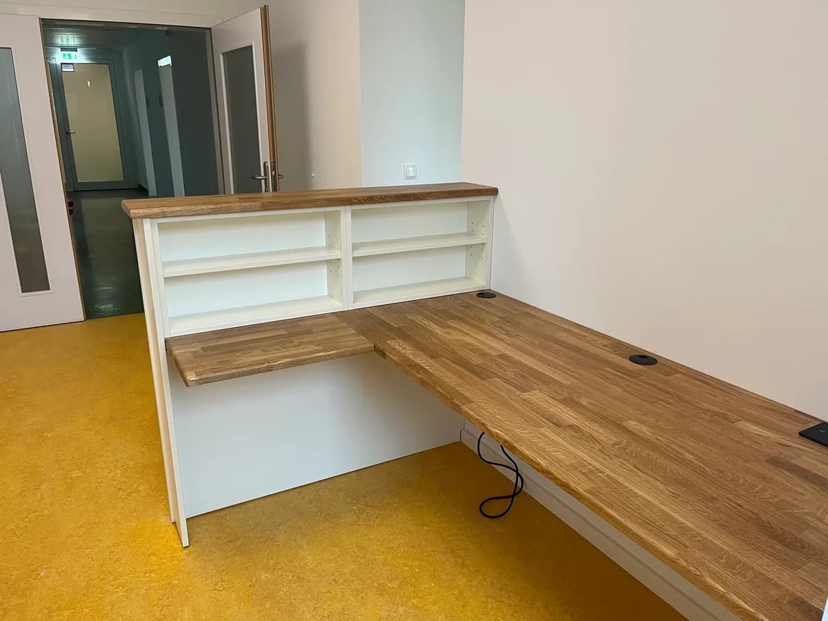 Der Tresen von der Raumseite: unter der Holzauflage zwei offene weiße Regalfächer, davor die tiefere Schreibplatte, die rechts an der Wand entlangläuft. Links eine offene Tür in einen Flur mit grünem Boden, im Raum gelbes Linoleum.