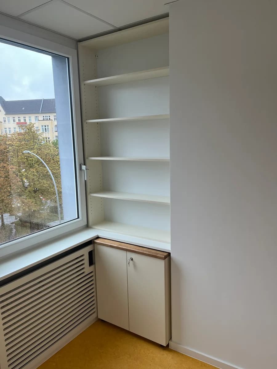 Das Einbauregal am Fenster: fünf weiße Fachböden über einem abschließbaren Unterschrank mit zwei Türen und heller Holzabdeckung. Links das Fenster mit Blick auf Straßenbäume, darunter ein verkleideter Heizkörper, gelbes Linoleum.