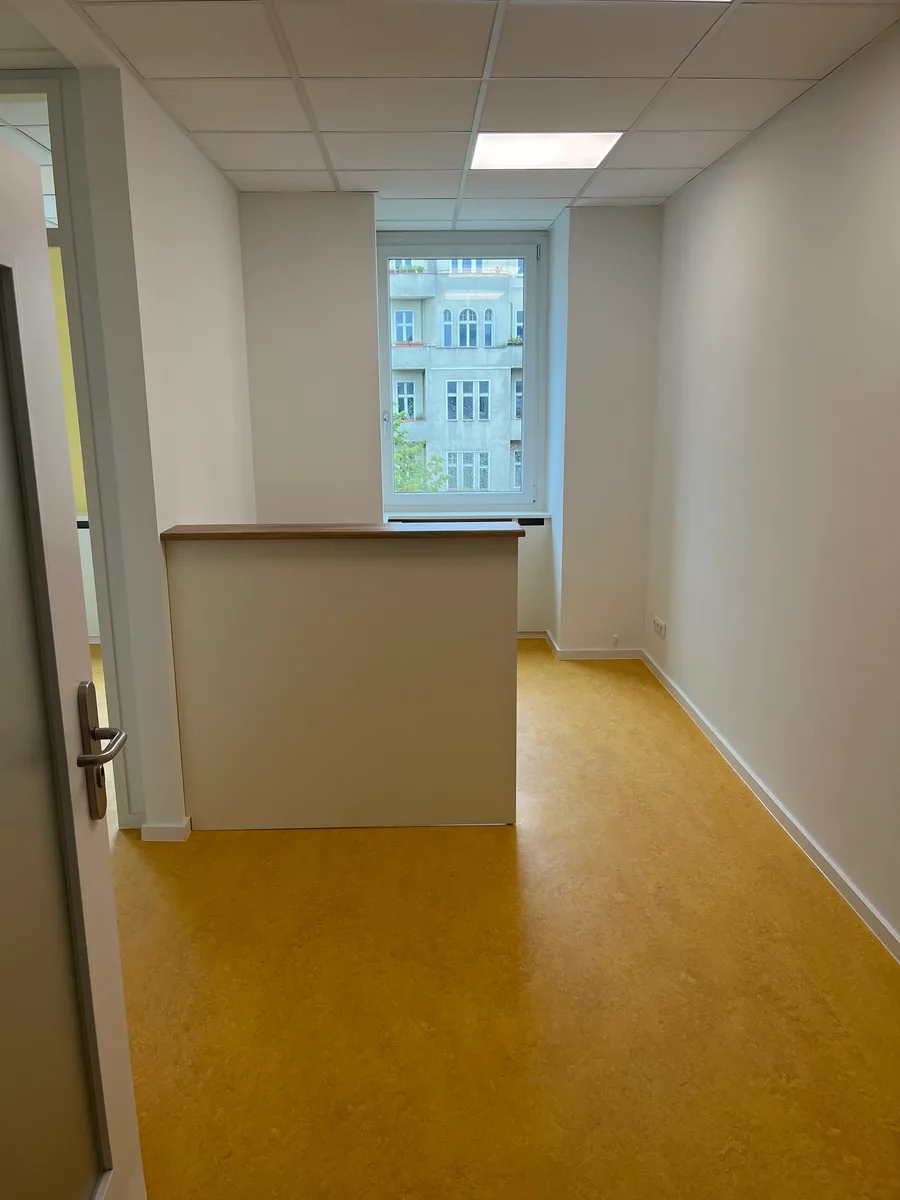 Der Raum von der Tür aus gesehen: der Tresen steht quer und schirmt den hinteren Teil mit dem Fenster ab. Eine Deckenleuchte, gelbes Linoleum, sonst ist der Raum leer.