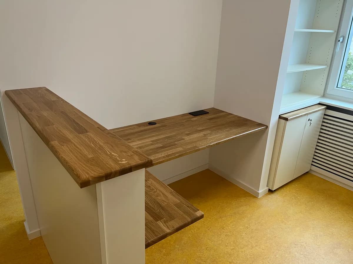 Der Empfangstresen von vorn, über Eck: eine niedrige weiße Brüstung mit durchgehender heller Holzauflage, die rechts in eine tiefere Schreibplatte mit schmaler Ablage darunter übergeht, in der Platte zwei runde Kabeldurchlässe. Dahinter ein weißes Regal über einem abschließbaren Unterschrank am Fenster, daneben ein verkleideter Heizkörper, gelbes Linoleum.