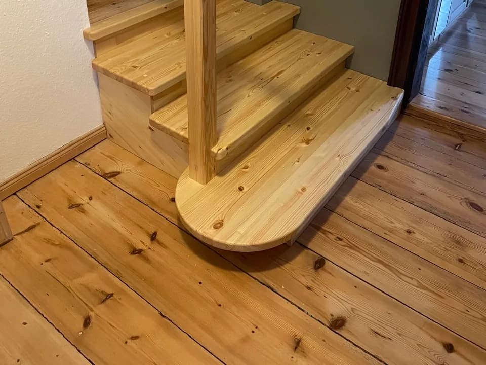 Podesttreppe aus geölter Kiefer mit drei Stufen, die unterste vorn abgerundet und weit vorgezogen. Der Handlauf sitzt auf einem eigenen Pfosten. Ringsum Dielenboden.