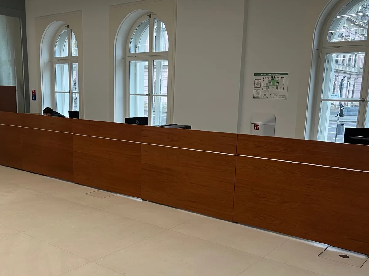 Durchlaufende Tischanlage aus furniertem Holz vor Rundbogenfenstern in einem Sitzungssaal.